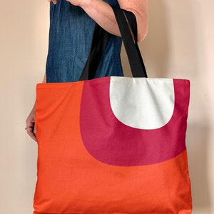 May include: A large orange tote bag with a red and white geometric design. The bag has black straps.