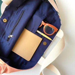 May include: A pink and green canvas tote bag with a navy blue lining. The bag has a zippered pocket and a small pocket on the inside. The bag is open and there is a notebook, a pen, and sunglasses inside.