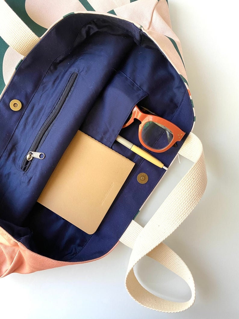 May include: A pink and green canvas tote bag with a navy blue lining. The bag has a zippered pocket and a small pocket on the inside. The bag is open and there is a notebook, a pen, and sunglasses inside.