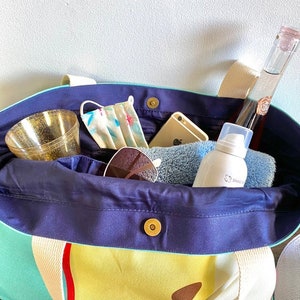 May include: A blue and green canvas tote bag with a yellow and red striped design. The bag is filled with various items, including a face mask, sunglasses, a phone, a bottle of liquid, and a tube of lotion.