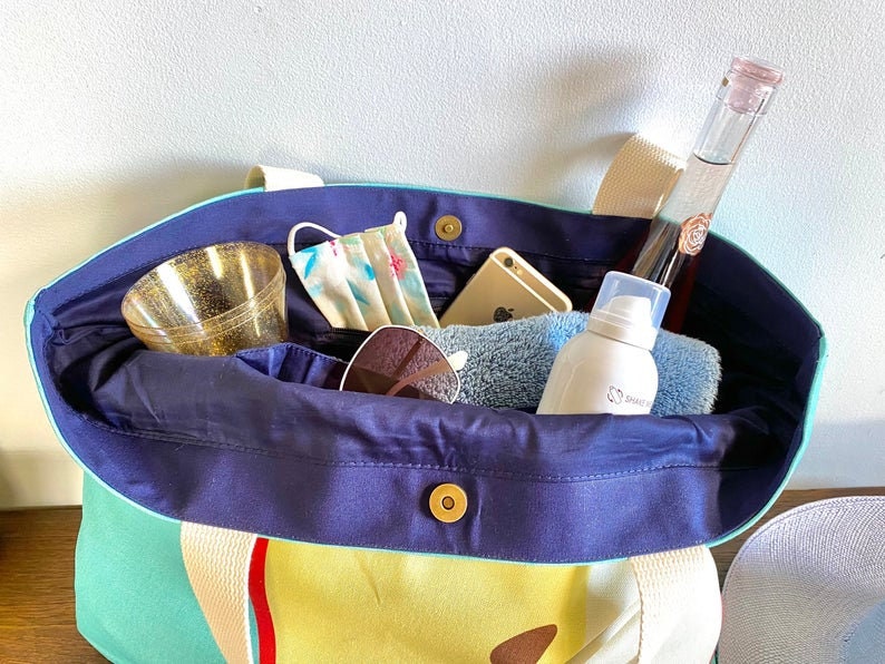 May include: A blue and green canvas tote bag with a yellow and red striped design. The bag is filled with various items, including a face mask, sunglasses, a phone, a bottle of liquid, and a tube of lotion.