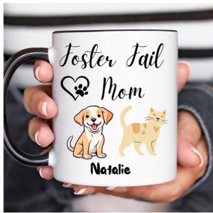 Peut inclure: Mug en céramique blanche avec un bord et une poignée noirs. Le mug a un cœur noir avec une empreinte de patte et le texte "Foster Fail Mom Natalie". Le mug présente également une image de dessin animé d'un chien et d'un chat.