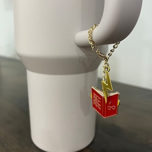 May include: A light gray tumbler with a gold chain charm. The charm features a red book with gold accents and a gold lightning bolt. The tumbler has a handle and a wide base.