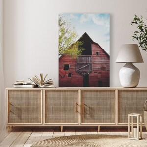 May include: A red barn with a green tree in front of it. The barn has a large door and a window. The tree is in the foreground of the image. The barn is in the background of the image. The image is a photograph.