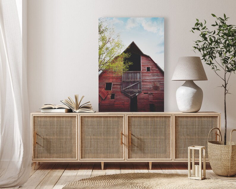 May include: A red barn with a green tree in front of it. The barn has a large door and a window. The tree is in the foreground of the image. The barn is in the background of the image. The image is a photograph.