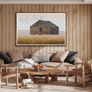 May include: A framed photograph of a weathered wooden cabin in a field with a flock of birds flying overhead. The cabin has a single window and a wooden door. The field is covered in tall grass.