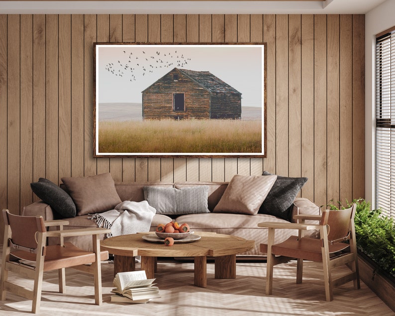 May include: A framed photograph of a weathered wooden cabin in a field with a flock of birds flying overhead. The cabin has a single window and a wooden door. The field is covered in tall grass.