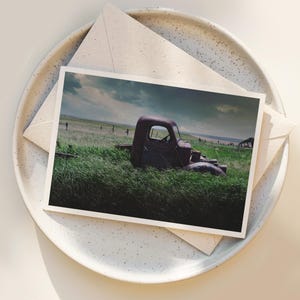 Pu&ograve; includere: Un camioncino rosso arrugginito &egrave; parzialmente immerso in un campo di erba verde alta. Il camioncino &egrave; rivolto verso il lato destro dell'immagine. Il cielo &egrave; blu nuvoloso con un tocco di arancione in lontananza.