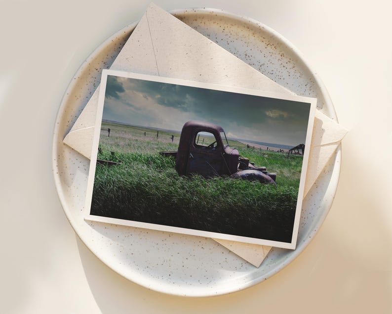 Pu&ograve; includere: Un camioncino rosso arrugginito &egrave; parzialmente immerso in un campo di erba verde alta. Il camioncino &egrave; rivolto verso il lato destro dell'immagine. Il cielo &egrave; blu nuvoloso con un tocco di arancione in lontananza.