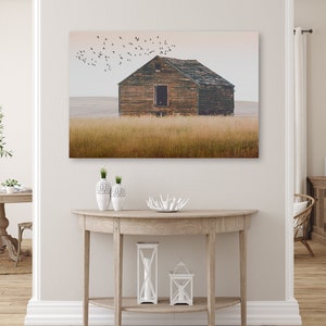 May include: A weathered wooden barn stands in a field of tall grass, with a flock of birds flying overhead. The sky is a soft, muted blue, and the barn is painted a dark green. The barn has a single window and a wooden door.