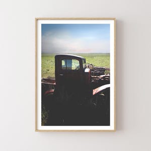 Puede incluir: Un cami&oacute;n vintage rojo oxidado se encuentra parcialmente sumergido en hierba verde alta. El cami&oacute;n est&aacute; mirando hacia el lado izquierdo de la imagen y la cabina es visible. El cami&oacute;n est&aacute; en un campo con un cielo azul en el fondo.