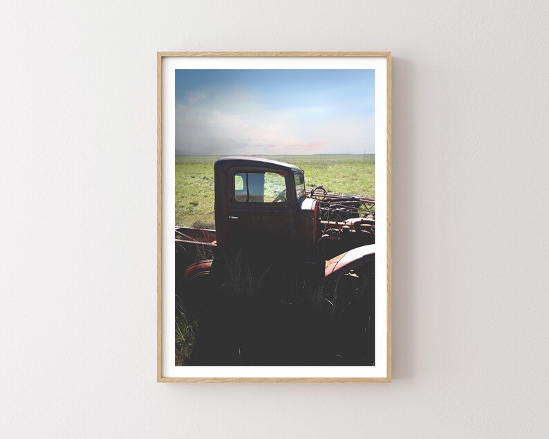 Puede incluir: Un cami&oacute;n vintage rojo oxidado se encuentra parcialmente sumergido en hierba verde alta. El cami&oacute;n est&aacute; mirando hacia el lado izquierdo de la imagen y la cabina es visible. El cami&oacute;n est&aacute; en un campo con un cielo azul en el fondo.