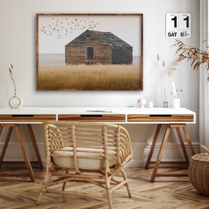 May include: A framed photograph of a weathered wooden house in a field with a flock of birds flying overhead. The house has a single window and a door. The field is covered in tall grass. The photograph is hanging on a white wall above a white desk with a wooden chair and a wicker basket.