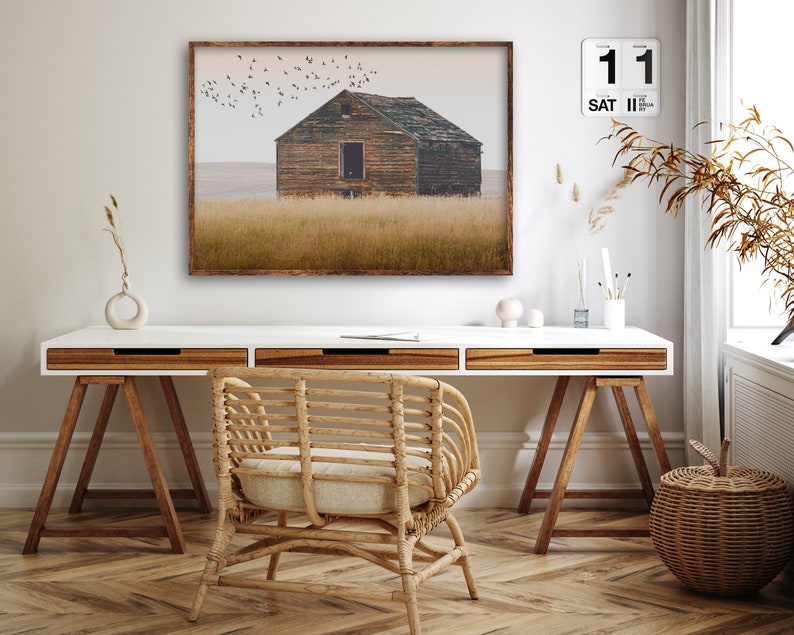 May include: A framed photograph of a weathered wooden house in a field with a flock of birds flying overhead. The house has a single window and a door. The field is covered in tall grass. The photograph is hanging on a white wall above a white desk with a wooden chair and a wicker basket.
