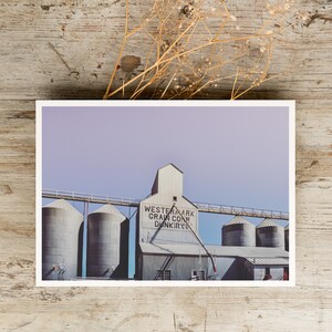 May include: A vintage photograph of a grain elevator with the text "Western Mark Grain Corp. Dunkirk" on the side. The building is made of metal and has a light gray color. The sky is a light purple color.
