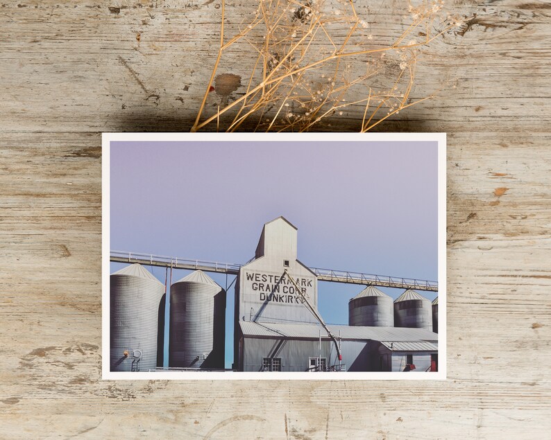 May include: A vintage photograph of a grain elevator with the text "Western Mark Grain Corp. Dunkirk" on the side. The building is made of metal and has a light gray color. The sky is a light purple color.
