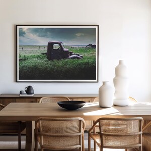 Pu&ograve; includere: Un camioncino rosso vintage parzialmente immerso in un'alta erba verde con un cielo tempestoso sullo sfondo. Il camioncino &egrave; parcheggiato in un campo con una recinzione di legno in lontananza. L'immagine &egrave; incorniciata e appesa a una parete bianca sopra un tavolo da pranzo in legno con sedie.