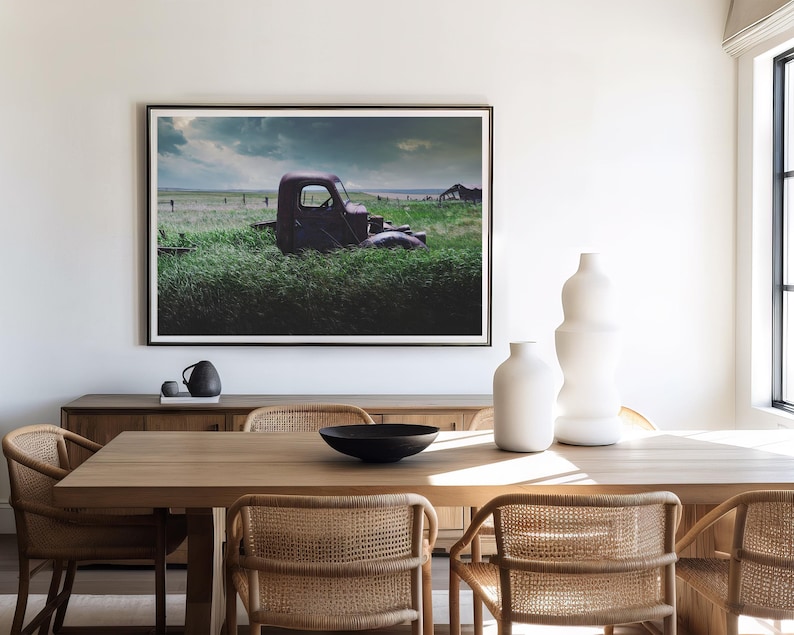 Pu&ograve; includere: Un camioncino rosso vintage parzialmente immerso in un'alta erba verde con un cielo tempestoso sullo sfondo. Il camioncino &egrave; parcheggiato in un campo con una recinzione di legno in lontananza. L'immagine &egrave; incorniciata e appesa a una parete bianca sopra un tavolo da pranzo in legno con sedie.