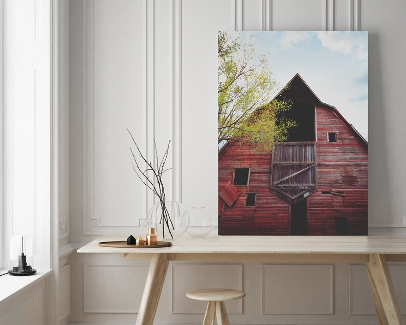 May include: A red barn with a large open doorway and a partially open door. The barn is weathered and has several windows. A tree with green leaves is in front of the barn.