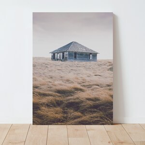 May include: A weathered wooden house stands alone in a field of tall, dry grass. The sky is overcast and the house appears abandoned.