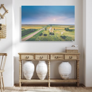 May include: An aerial view of a white church with a steeple in a field with a road leading to it. The church is surrounded by a cemetery with headstones. The sky is blue with clouds and the sun is shining.