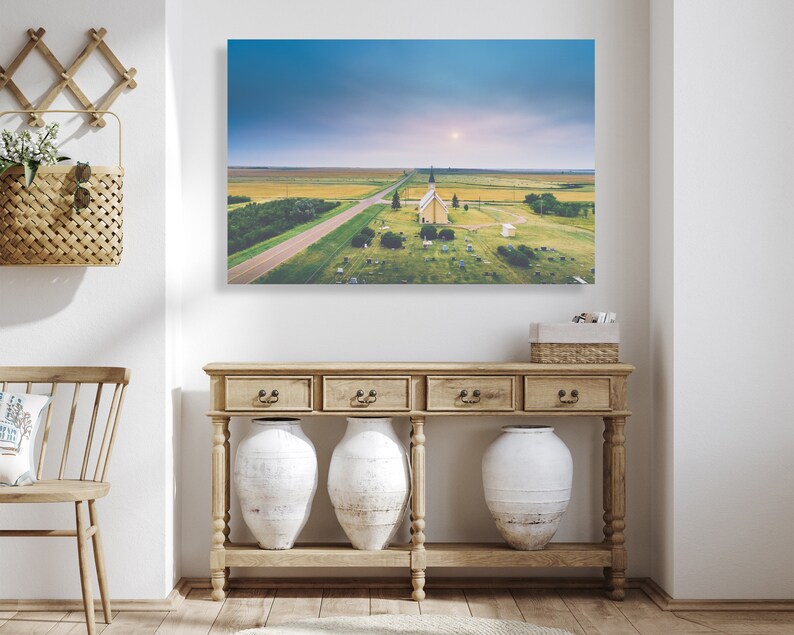 May include: An aerial view of a white church with a steeple in a field with a road leading to it. The church is surrounded by a cemetery with headstones. The sky is blue with clouds and the sun is shining.