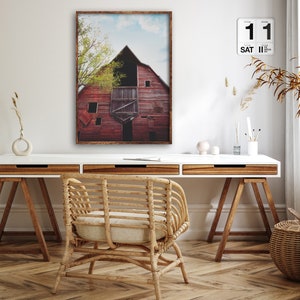 May include: A framed photograph of a red barn with a blue sky and green trees in the background. The barn is weathered and has a rustic charm. The photo is hanging on a white wall above a white desk with a wooden chair and a wicker basket.