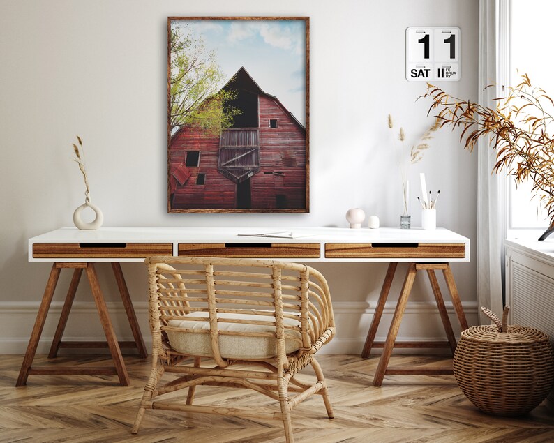 May include: A framed photograph of a red barn with a blue sky and green trees in the background. The barn is weathered and has a rustic charm. The photo is hanging on a white wall above a white desk with a wooden chair and a wicker basket.