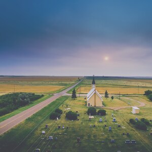 May include: An aerial view of a small, yellow church with a black spire, surrounded by a grassy graveyard. A paved road leads to the church, and a field of golden wheat stretches out in the distance.
