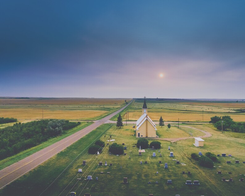 May include: An aerial view of a small, yellow church with a black spire, surrounded by a grassy graveyard. A paved road leads to the church, and a field of golden wheat stretches out in the distance.