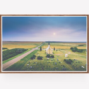 May include: An aerial view of a small, white church with a spire, surrounded by a grassy graveyard. The church is situated in a rural area with a long, straight road leading to it. The sky is a soft blue with a hint of pink.