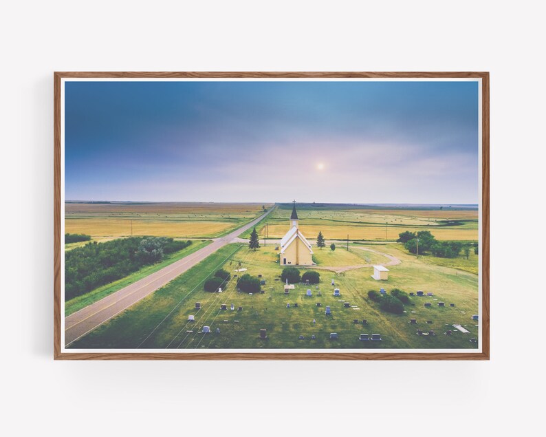 May include: An aerial view of a small, white church with a spire, surrounded by a grassy graveyard. The church is situated in a rural area with a long, straight road leading to it. The sky is a soft blue with a hint of pink.