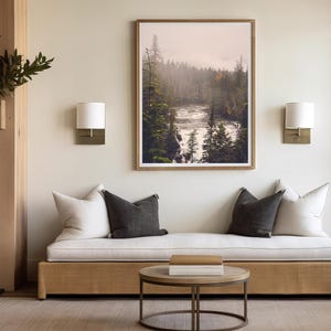 May include: A white couch with two pillows, one white and one gray, sits in front of a fireplace and a framed photograph of a misty forest scene with a river flowing through it.