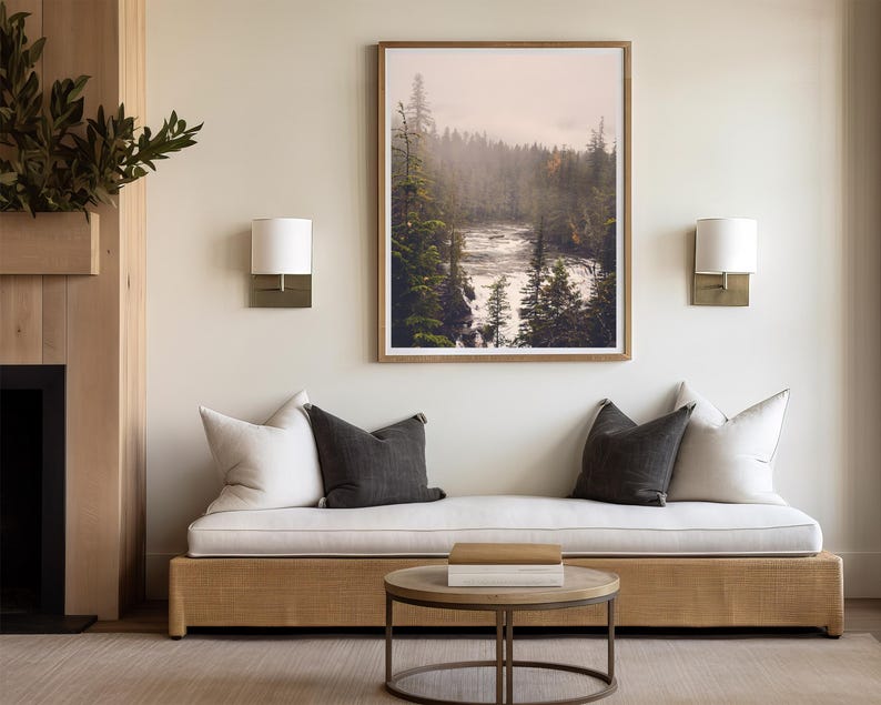 May include: A white couch with two pillows, one white and one gray, sits in front of a fireplace and a framed photograph of a misty forest scene with a river flowing through it.