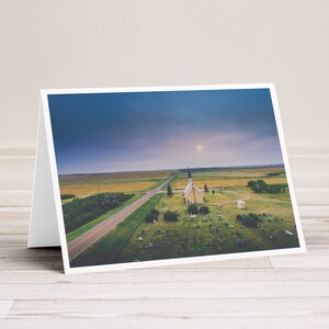 May include: An aerial view of a small, white church with a spire, surrounded by a grassy graveyard. The church is situated in a rural area with a long, straight road leading to it. The sky is a soft blue with a few clouds.