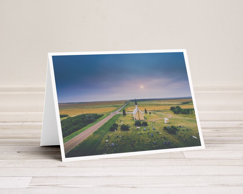 May include: An aerial view of a small, white church with a spire, surrounded by a grassy graveyard. The church is situated in a rural area with a long, straight road leading to it. The sky is a soft blue with a few clouds.