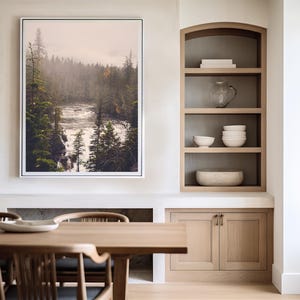 May include: A framed photograph of a misty forest scene with a river flowing through it. The photograph is hanging on a white wall above a wooden table and cabinets.