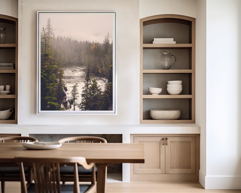 May include: A framed photograph of a misty forest scene with a river flowing through it. The photograph is hanging on a white wall above a wooden table and cabinets.