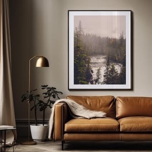May include: A framed photograph of a misty forest scene with a river flowing through it. The photograph is in a brown frame and is hanging on a wall.