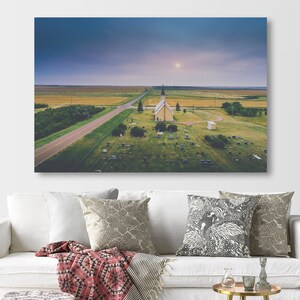 May include: An aerial view of a small, white church with a spire, surrounded by a grassy graveyard. The church is situated in a rural area with a long, straight road leading to it. The sky is a soft blue with a few clouds.