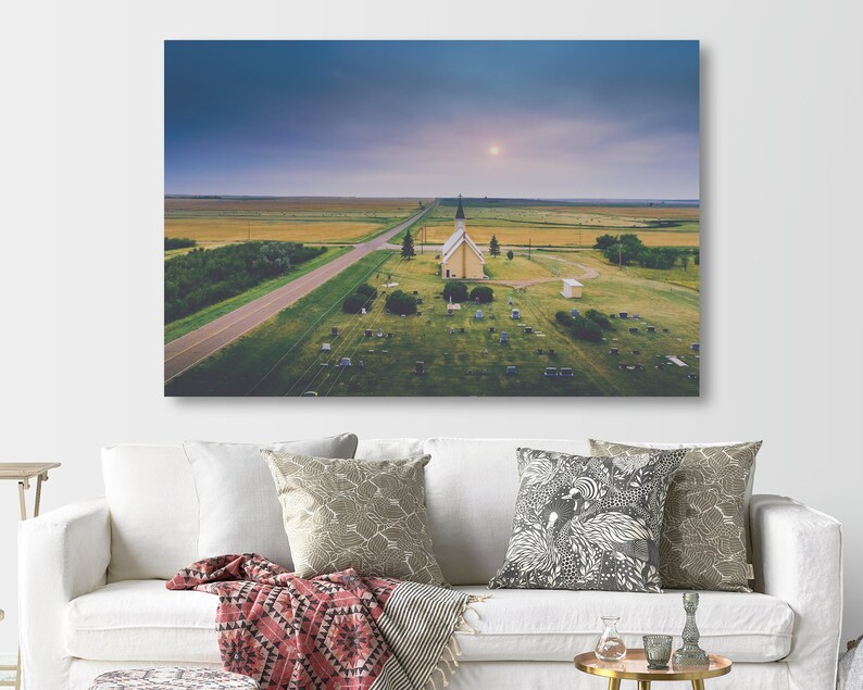 May include: An aerial view of a small, white church with a spire, surrounded by a grassy graveyard. The church is situated in a rural area with a long, straight road leading to it. The sky is a soft blue with a few clouds.