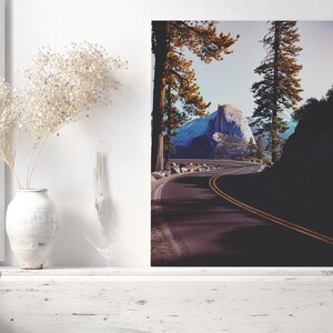 Pu&ograve; includere: Una strada tortuosa conduce a una vista di Half Dome, una formazione rocciosa di granito nel Parco Nazionale di Yosemite, in California. La strada &egrave; fiancheggiata da alberi alti e il cielo &egrave; di un blu brillante.