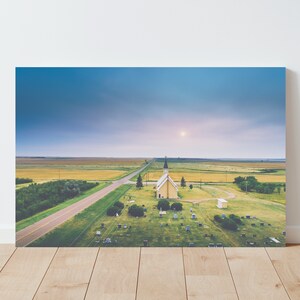 May include: An aerial view of a small, yellow church with a white spire, surrounded by a graveyard. The church is situated in a rural area with a long, straight road leading to it. The sky is a soft blue with a few clouds.