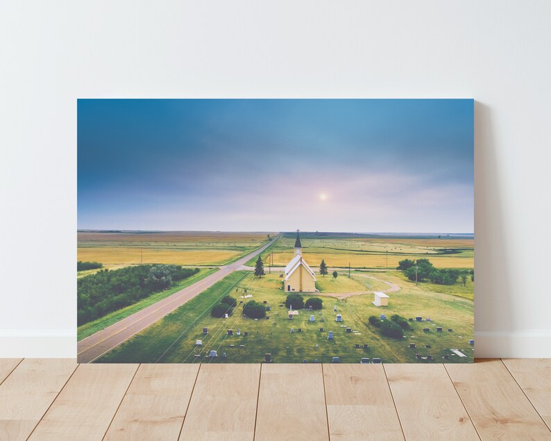 May include: An aerial view of a small, yellow church with a white spire, surrounded by a graveyard. The church is situated in a rural area with a long, straight road leading to it. The sky is a soft blue with a few clouds.