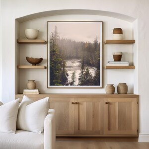 May include: A living room interior with a white couch, two white pillows, and a wooden cabinet with three doors. The cabinet is decorated with two brown ceramic vases and a framed print of a misty forest scene with a river.