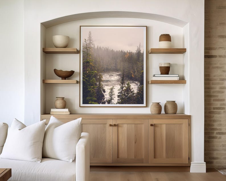 May include: A living room interior with a white couch, two white pillows, and a wooden cabinet with three doors. The cabinet is decorated with two brown ceramic vases and a framed print of a misty forest scene with a river.