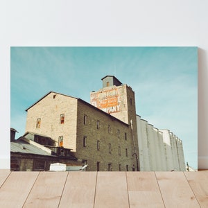 Peut inclure: Une photographie vintage d'un grand bâtiment en brique avec une enseigne fanée qui indique "North Dakota Mill & Elevator Company". Le bâtiment est doté d'une série de silos blancs et hauts qui y sont attachés.