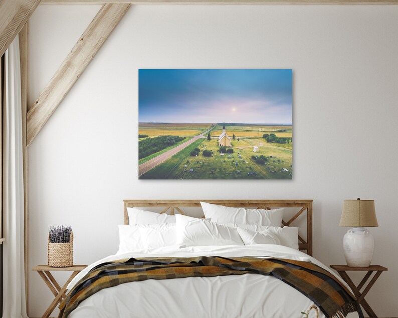 May include: An aerial view of a white church with a steeple, surrounded by a grassy field and a long, straight road leading to it. The sky is a soft blue with a few clouds.