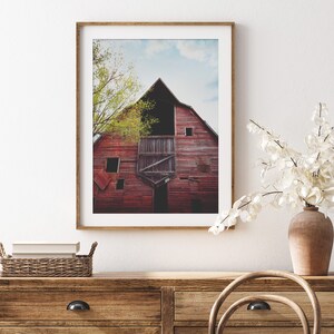 May include: A framed photograph of a red barn with a wooden door and a window. The barn is surrounded by green trees and a blue sky with white clouds.