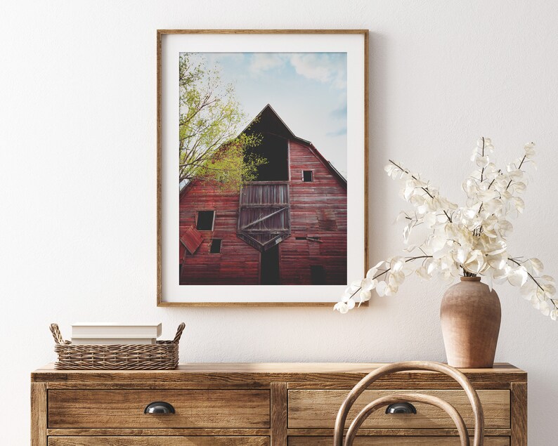 May include: A framed photograph of a red barn with a wooden door and a window. The barn is surrounded by green trees and a blue sky with white clouds.